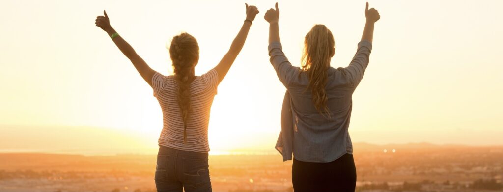 Two girls standing, arms raised, watching the sunset